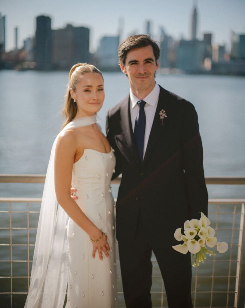 beautiful bride and groom pose waterside with the city stretching behind them, the classic hairstyle is a 2026 hair trend brides are loving!