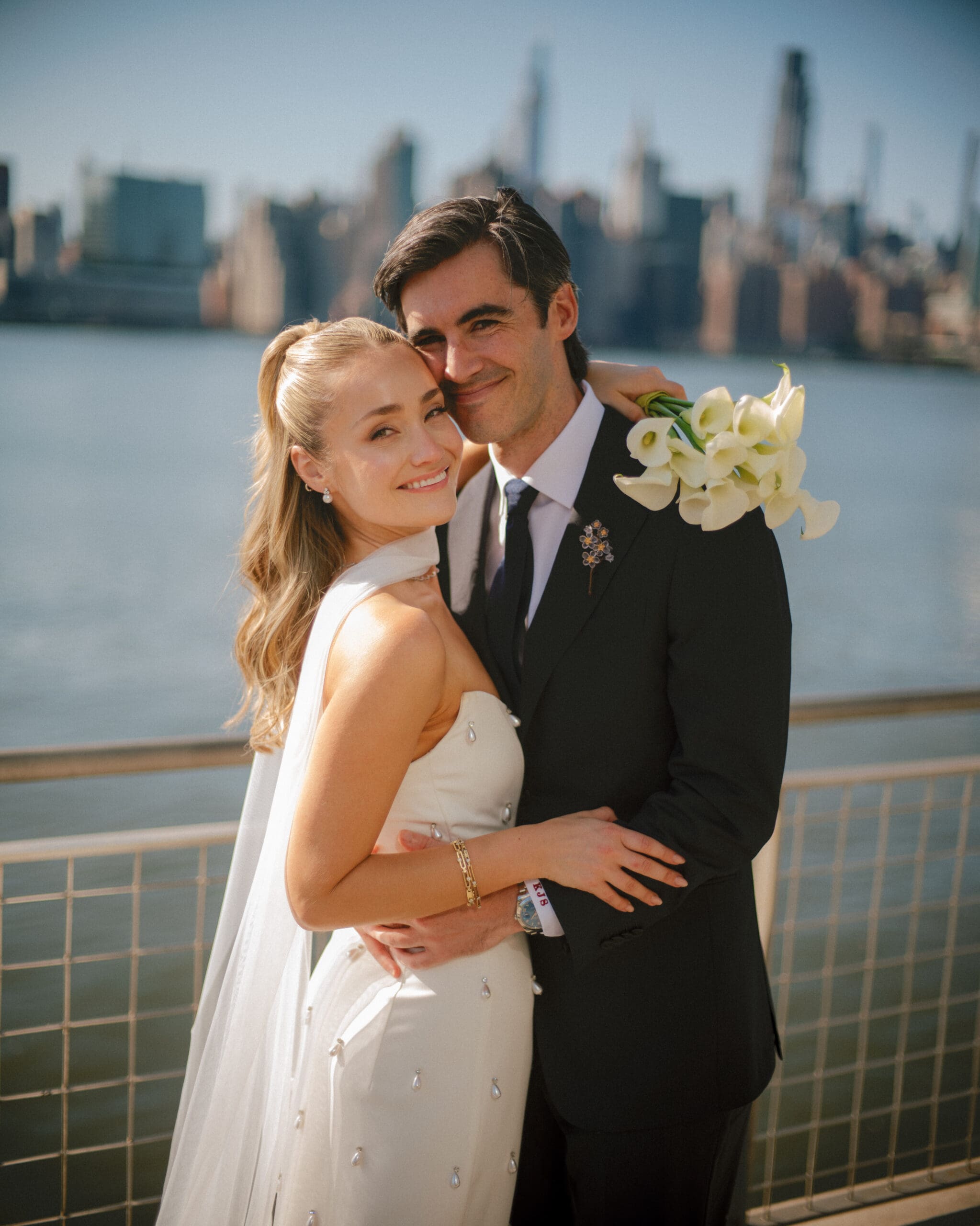 beautiful bride and groom pose waterside with the city stretching behind them, the classic hairstyle is a 2026 hair trend brides are loving!